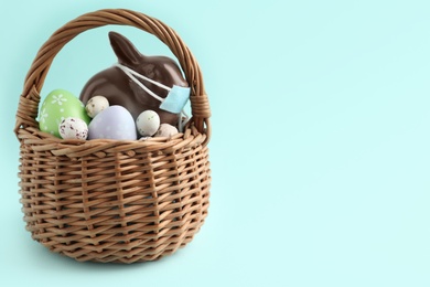 Photo of Chocolate bunny with protective mask and eggs in basket on light blue background, space for text. Easter holiday during COVID-19 quarantine