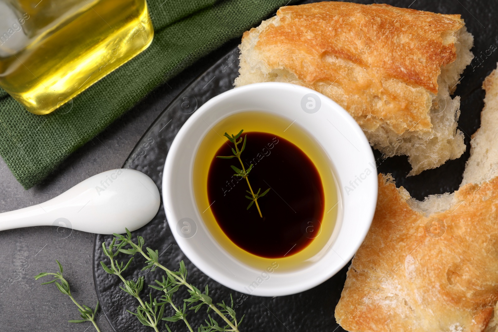 Photo of Bowl of organic balsamic vinegar with oil, thyme and bread on grey table, flat lay