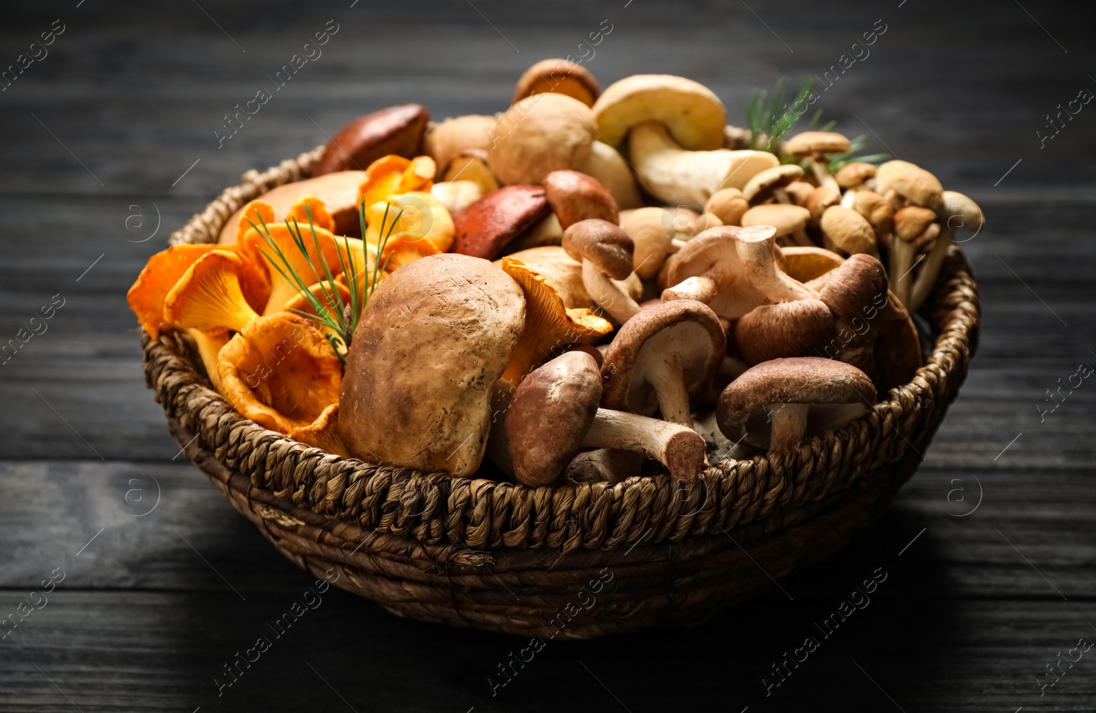 Photo of Different fresh wild mushrooms in wicker bowl on black wooden table, top view