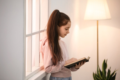 Photo of Cute little girl reading book near window at home