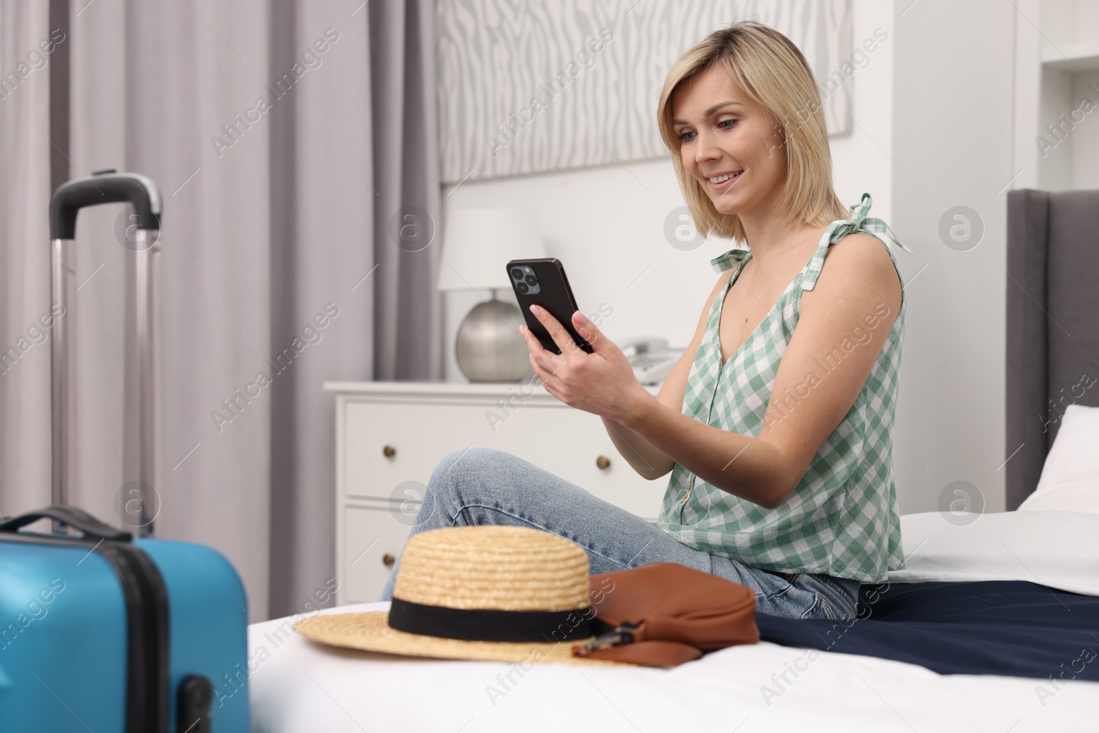 Photo of Smiling guest with smartphone relaxing on bed in stylish hotel room