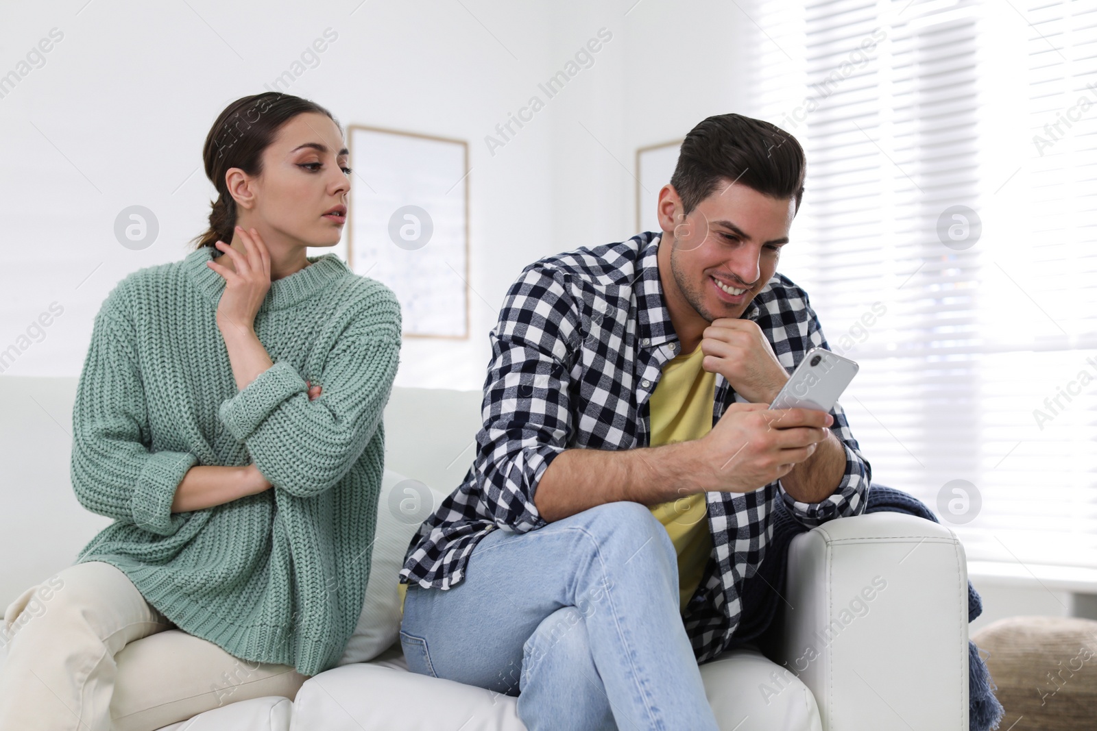 Photo of Distrustful woman peering into boyfriend's smartphone at home. Jealousy in relationship