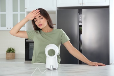 Photo of Woman with portable fan suffering from heat in kitchen. Summer season