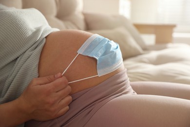 Photo of Young pregnant woman with medical mask on her belly indoors, closeup