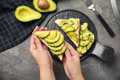 Photo of Woman holding tasty avocado toast over grey table, top view