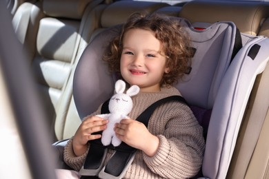 Photo of Cute little girl with toy rabbit sitting in child safety seat inside car