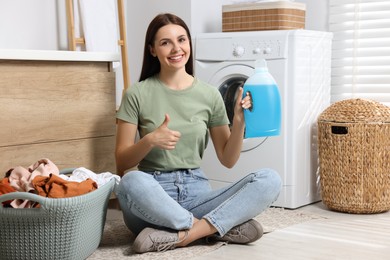 Woman sitting on floor near washing machine, holding fabric softener and showing thumbs up in bathroom