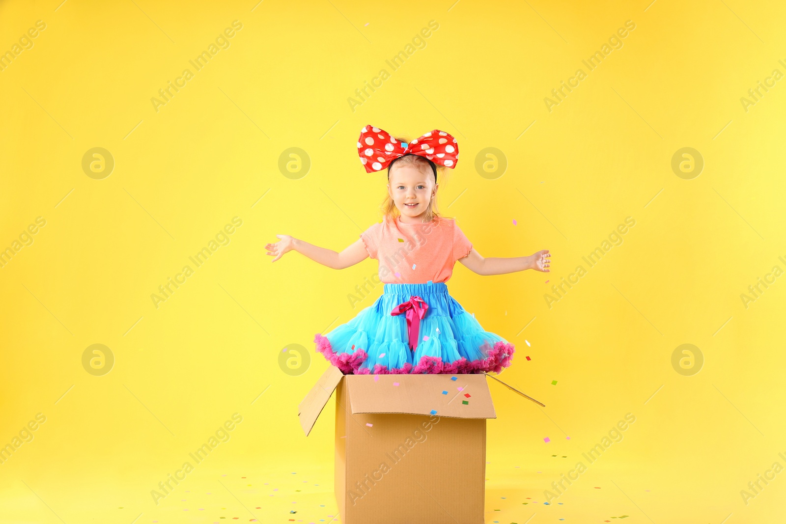 Photo of Little girl with large bow in cardboard box under confetti shower on yellow background. April fool's day