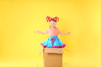 Little girl with large bow in cardboard box under confetti shower on yellow background. April fool's day