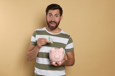 Photo of Excited man putting money into piggy bank on beige background