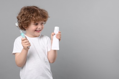 Cute little boy holding electric toothbrush and tube of toothpaste on light grey background, space for text