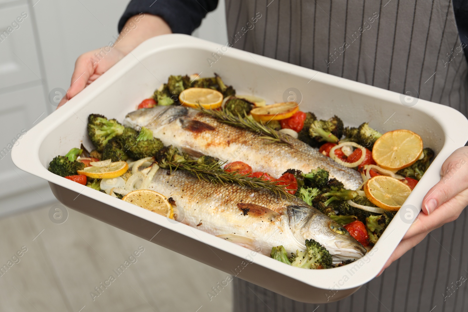 Photo of Woman holding baking dish with delicious fish and vegetables indoors, closeup