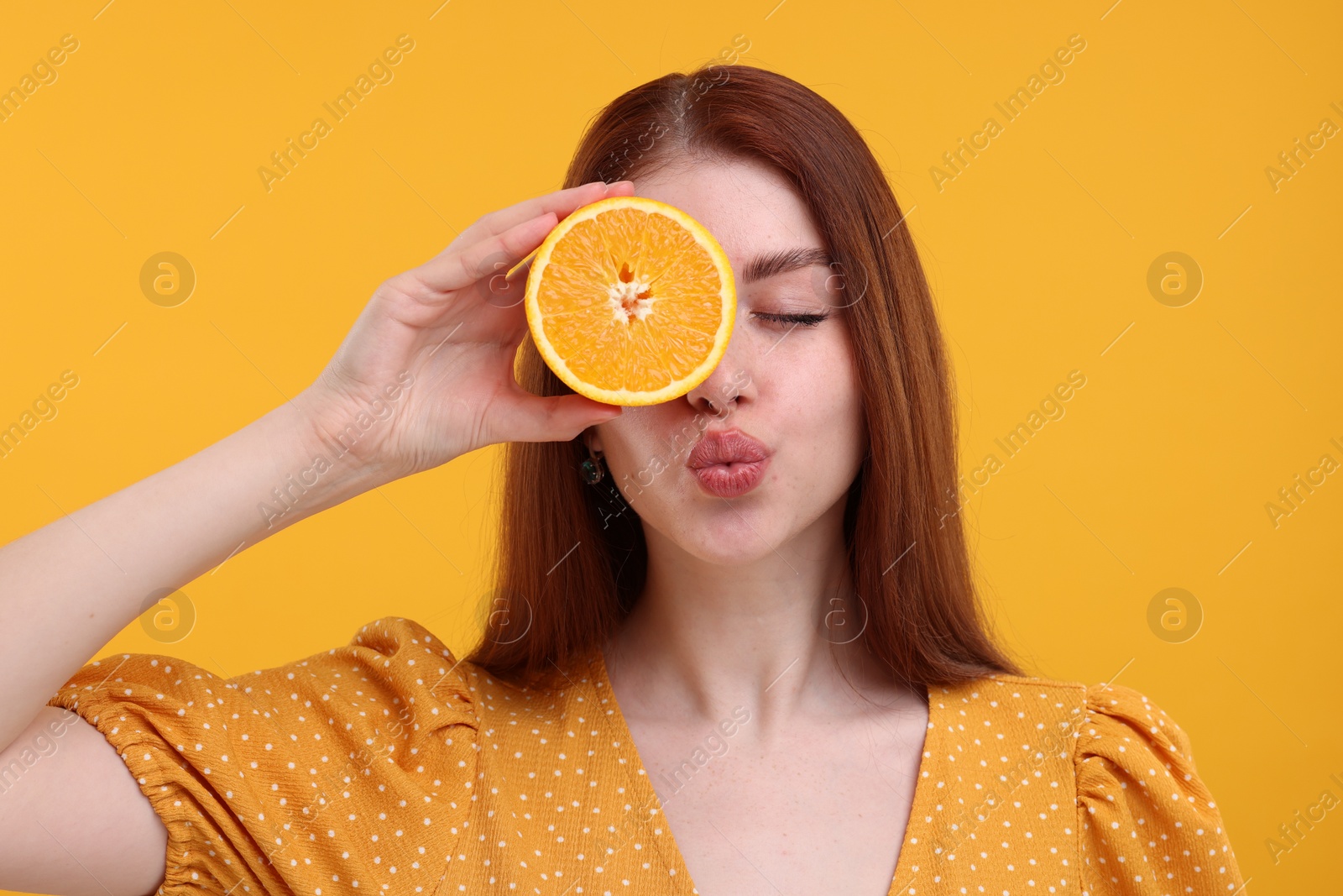 Photo of Beautiful woman covering eye with half of orange and sending air kiss on yellow background