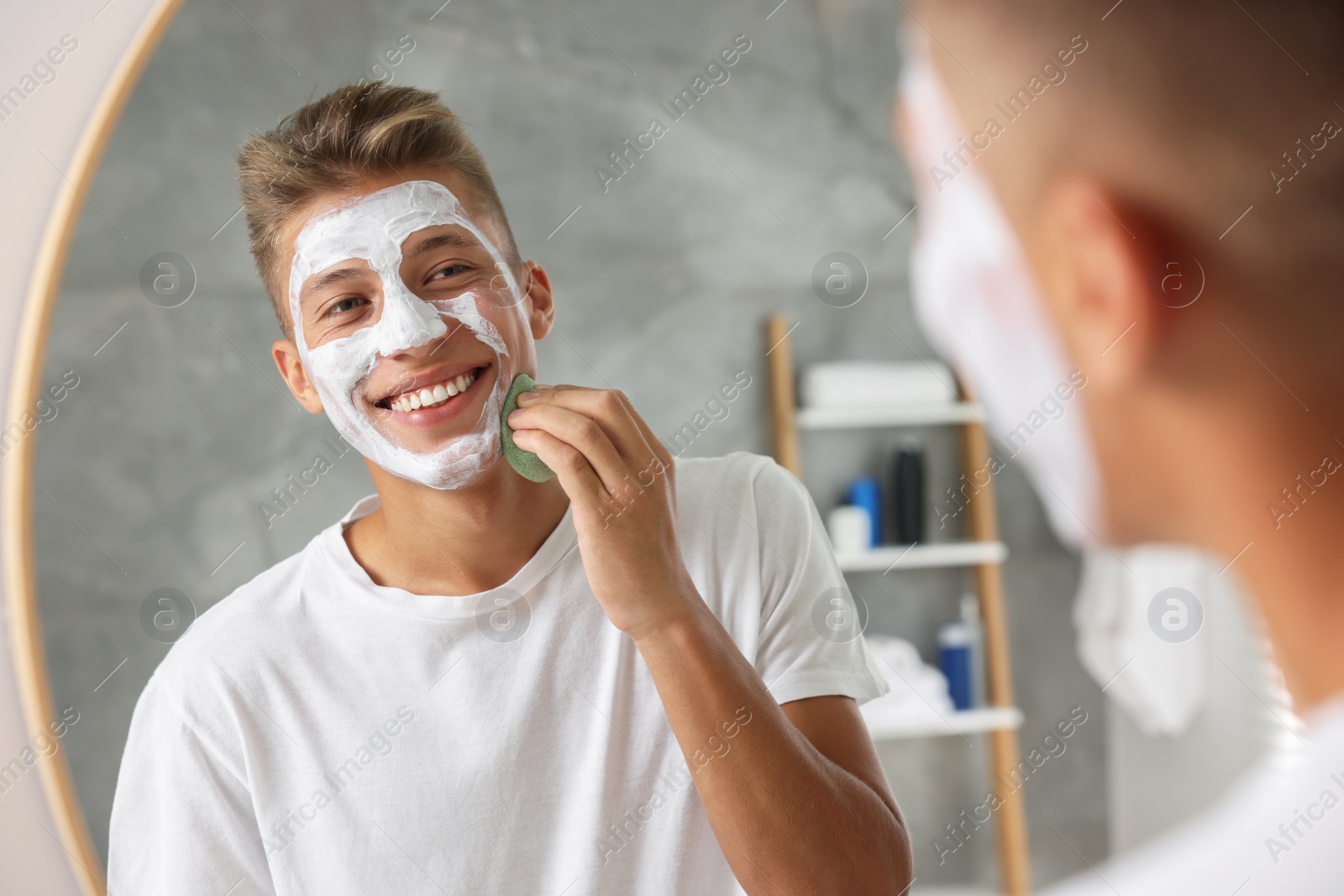 Photo of Happy young man washing off face mask with sponge near mirror in bathroom