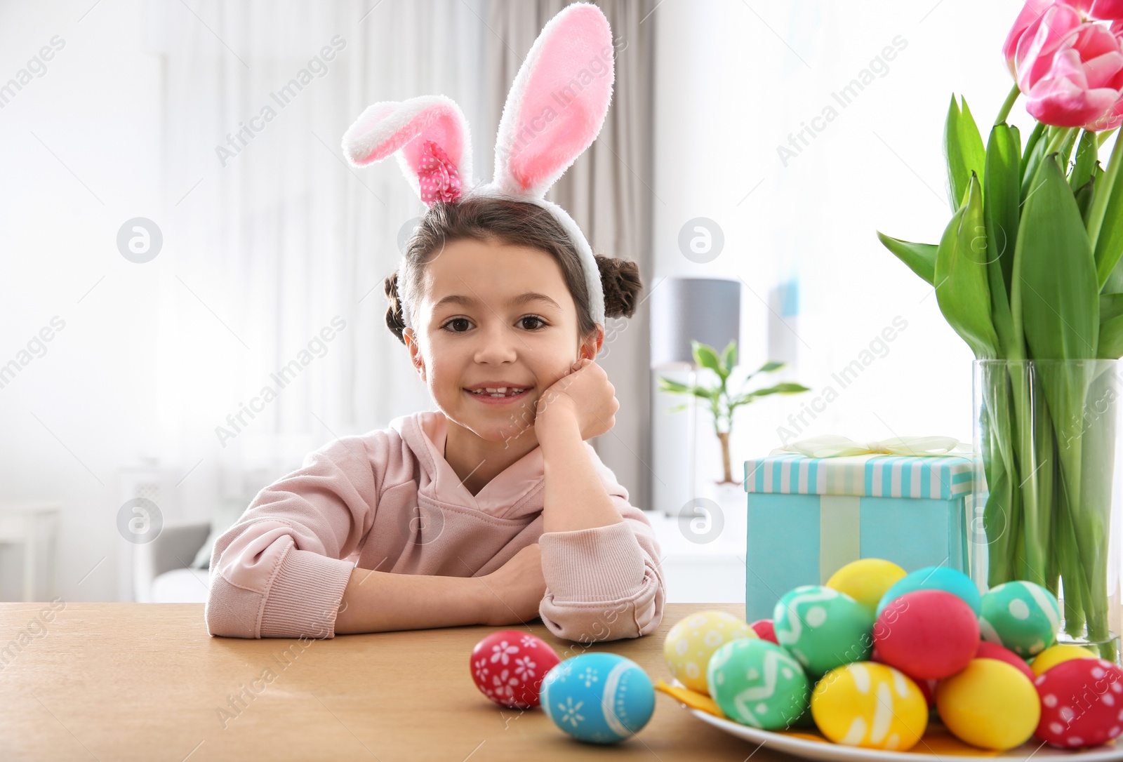 Photo of Cute little girl with bunny ears headband and painted Easter eggs sitting at table in room