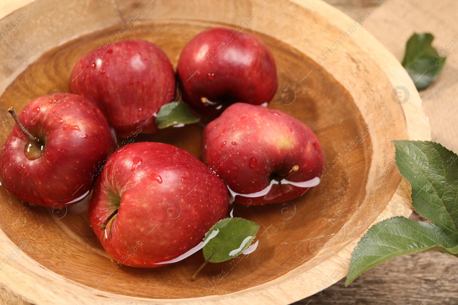 Photo of Fresh red apples in bowl with water and leaves on wooden table, closeup