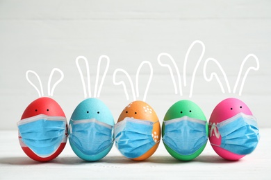 Image of COVID-19 pandemic. Bright Easter eggs with cute bunny ears in protective masks on white table