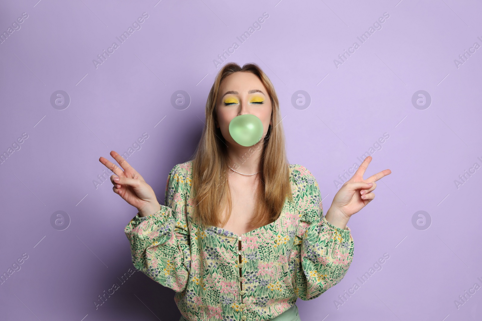 Photo of Fashionable young woman with bright makeup blowing bubblegum on lilac background