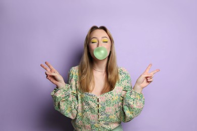 Fashionable young woman with bright makeup blowing bubblegum on lilac background