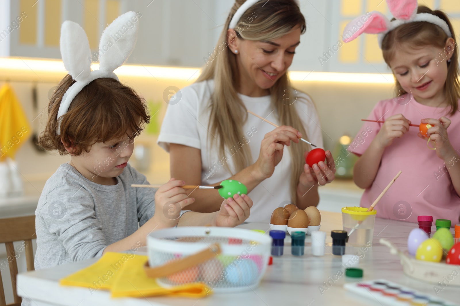 Photo of Easter celebration. Happy family with bunny ears painting eggs at white marble table in kitchen