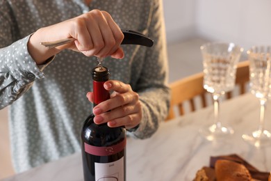 Romantic dinner. Woman opening wine bottle with corkscrew at table indoors, closeup
