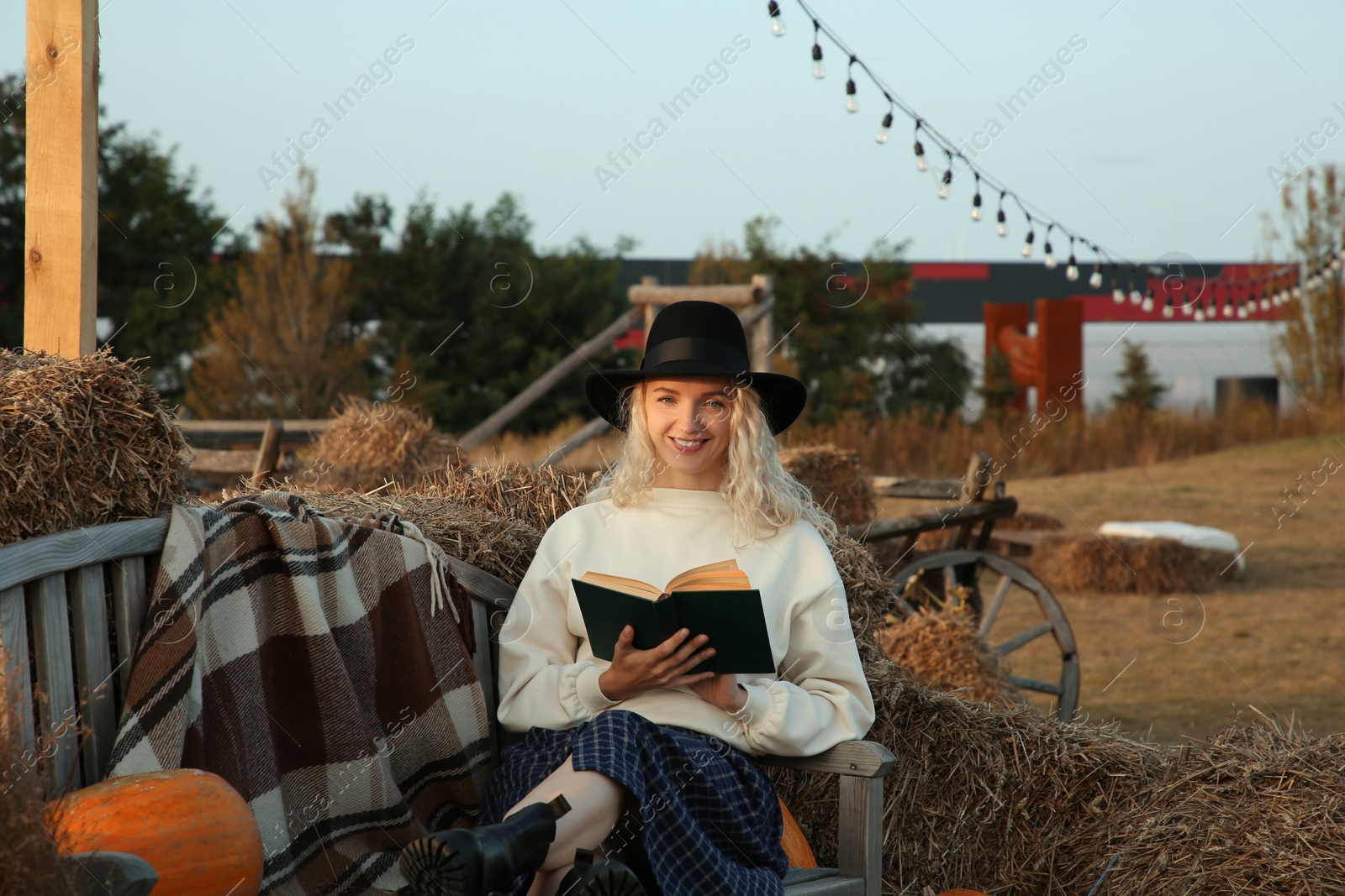 Photo of Beautiful woman with book sitting on wooden bench near hay bales outdoors. Autumn season