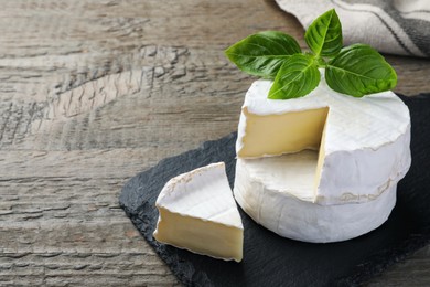 Tasty cut and whole brie cheeses with basil on wooden table, space for text