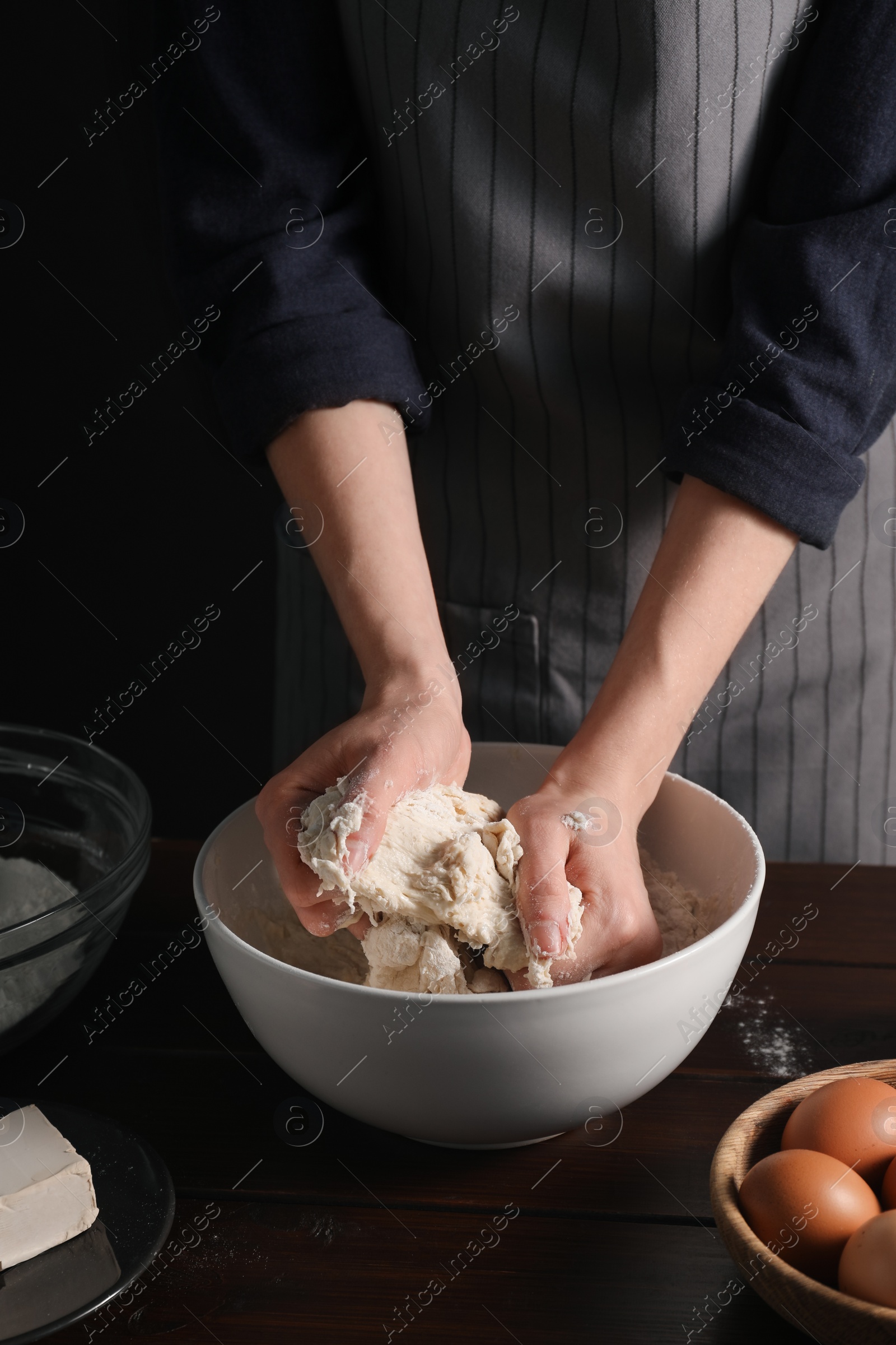 Photo of Making bread. Woman kneading dough at wooden table on dark background, closeup