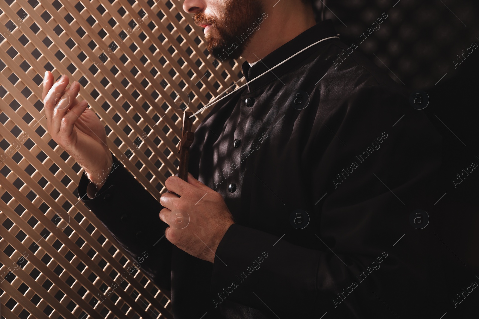 Photo of Catholic priest in cassock holding cross and talking to parishioner in confessional, closeup