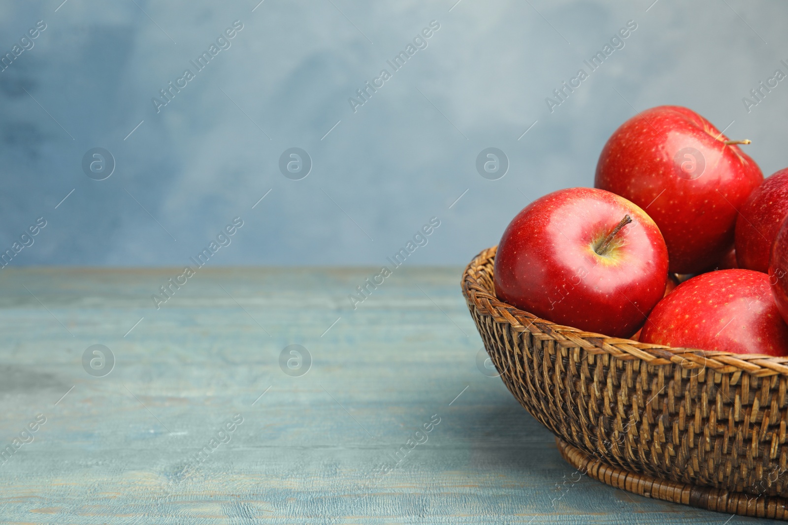 Photo of Wicker bowl with ripe juicy red apples on wooden table against blue background. Space for text