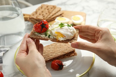 Woman eating fresh rye crispbread with poached egg, cream cheese and grilled tomato at table, closeup