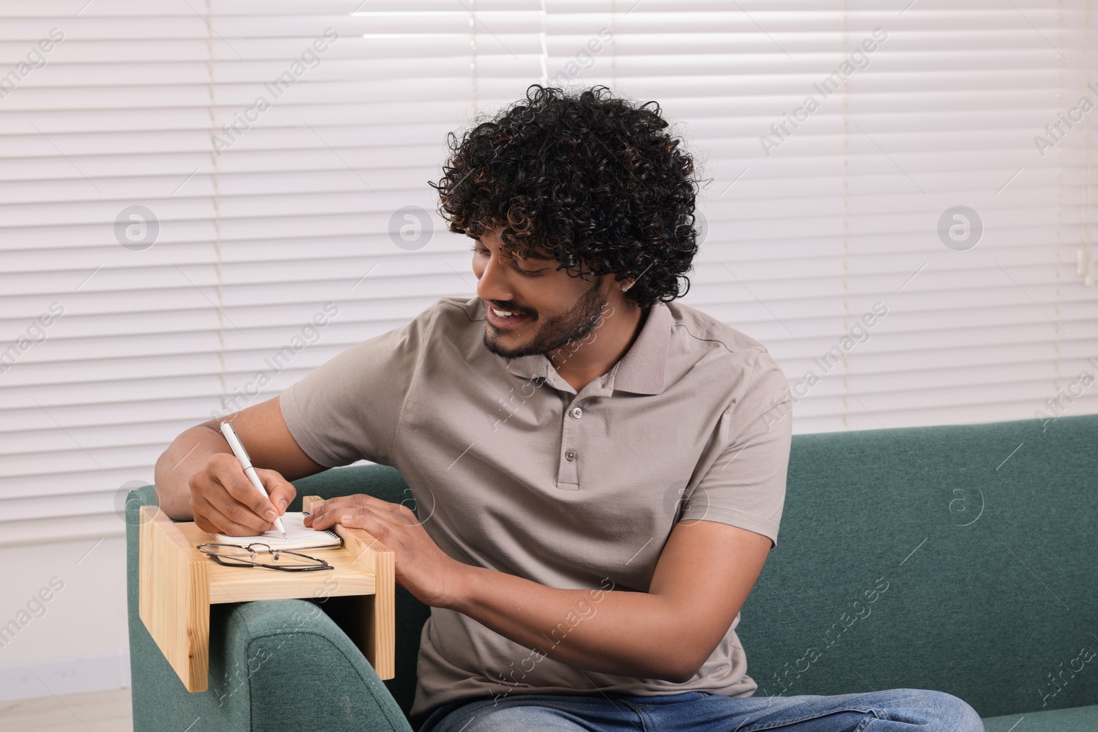 Photo of Happy man writing in notebook on sofa armrest wooden table at home