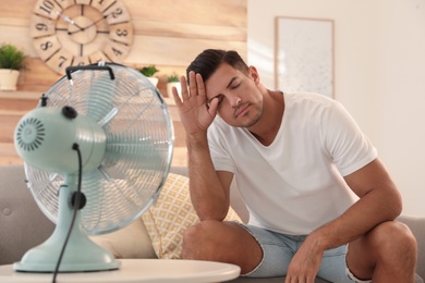 Photo of Man enjoying air flow from fan on sofa in living room. Summer heat