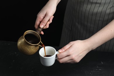 Photo of Turkish coffee. Woman pouring brewed beverage from cezve into cup at black table, closeup