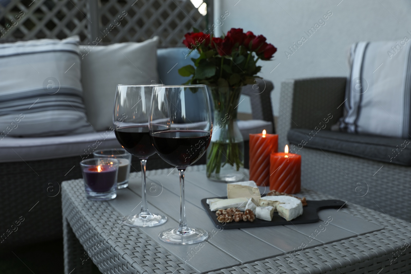 Photo of Glasses of wine, vase with roses, burning candles and snacks on rattan table at balcony in evening, closeup