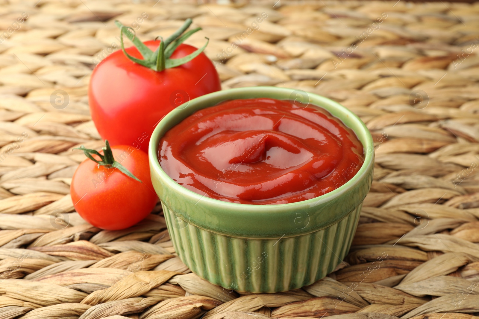 Photo of Bowl of tasty ketchup and tomatoes on wicker mat, closeup