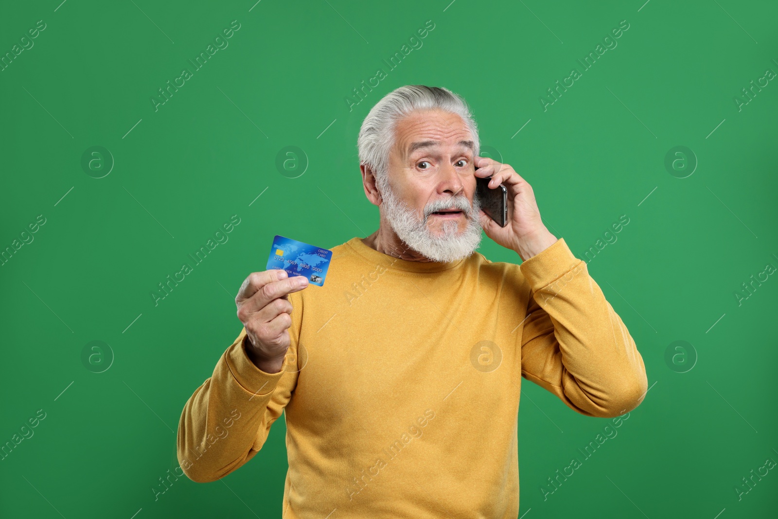 Photo of Confused senior man with credit card talking on smartphone against green background. Be careful - fraud