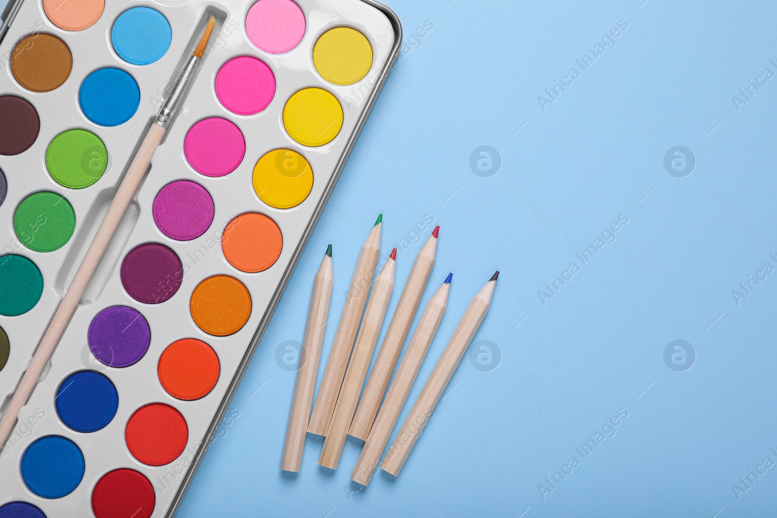 Photo of Watercolor palette with brush and colorful pencils on light blue background, flat lay. Space for text