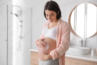 Photo of Young pregnant woman applying cosmetic product on belly in bathroom