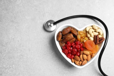 Photo of Heart shaped bowl with nuts and dried fruits near stethoscope on grey background, top view. Space for text
