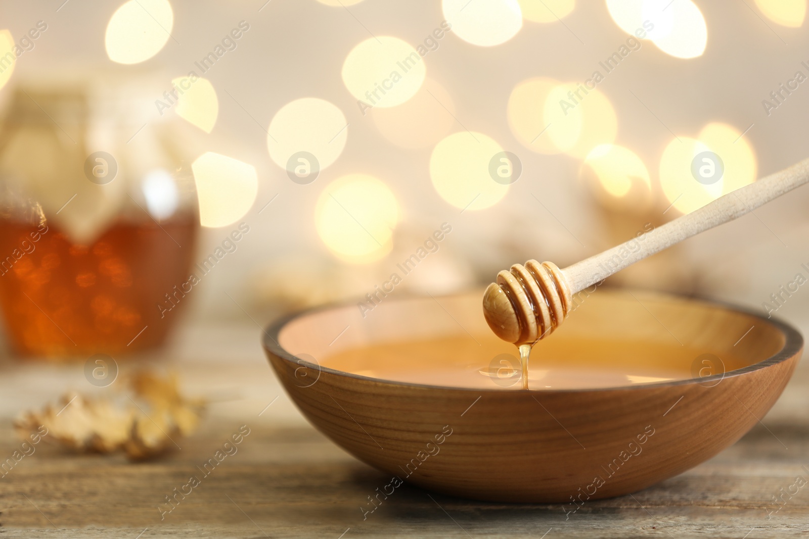 Photo of Honey dripping from dipper into bowl on table against blurred lights. Space for text