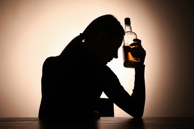 Photo of Alcohol addiction. Silhouette of woman with bottle of whiskey at wooden table, backlit