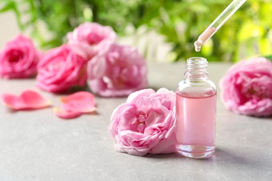 Photo of Dripping rose essential oil into bottle with pipette and fresh flowers on grey table, space for text