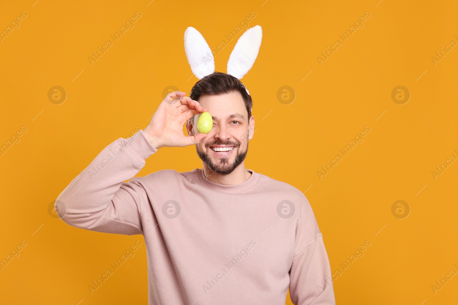 Photo of Happy man in bunny ears headband holding painted Easter egg on orange background