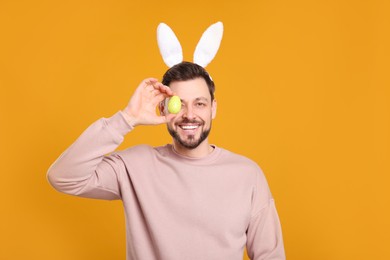 Photo of Happy man in bunny ears headband holding painted Easter egg on orange background