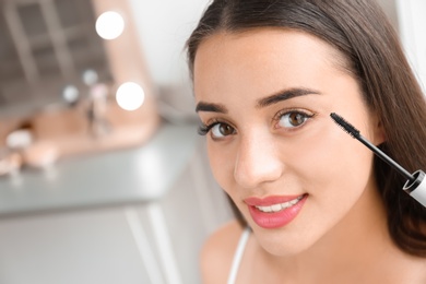 Photo of Attractive young woman applying mascara on her eyelashes against blurred background