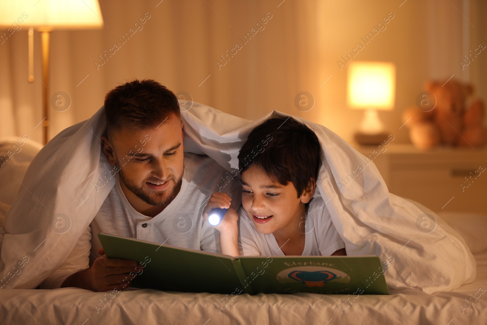 Photo of Father and son with flashlight reading book under blanket at home