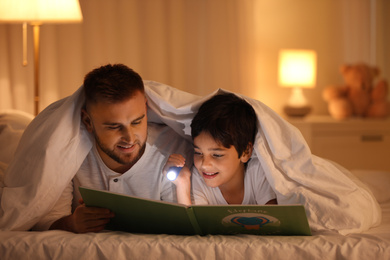 Photo of Father and son with flashlight reading book under blanket at home