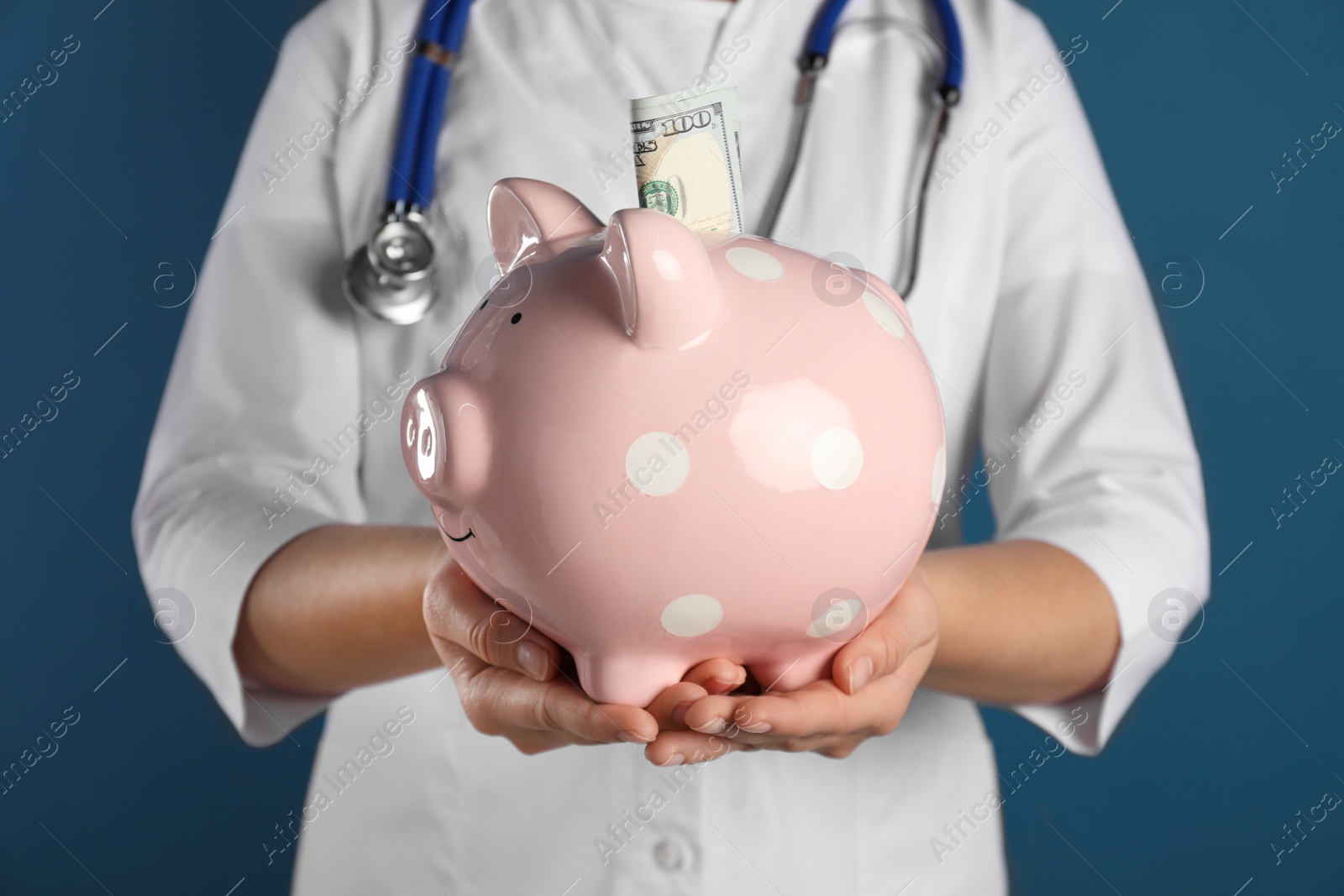 Photo of Doctor holding piggy bank with money on blue background, closeup. Medical insurance