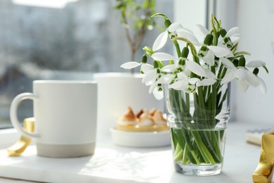 Photo of Beautiful snowdrops, cup of drink and dessert on white tray near window. Space for text